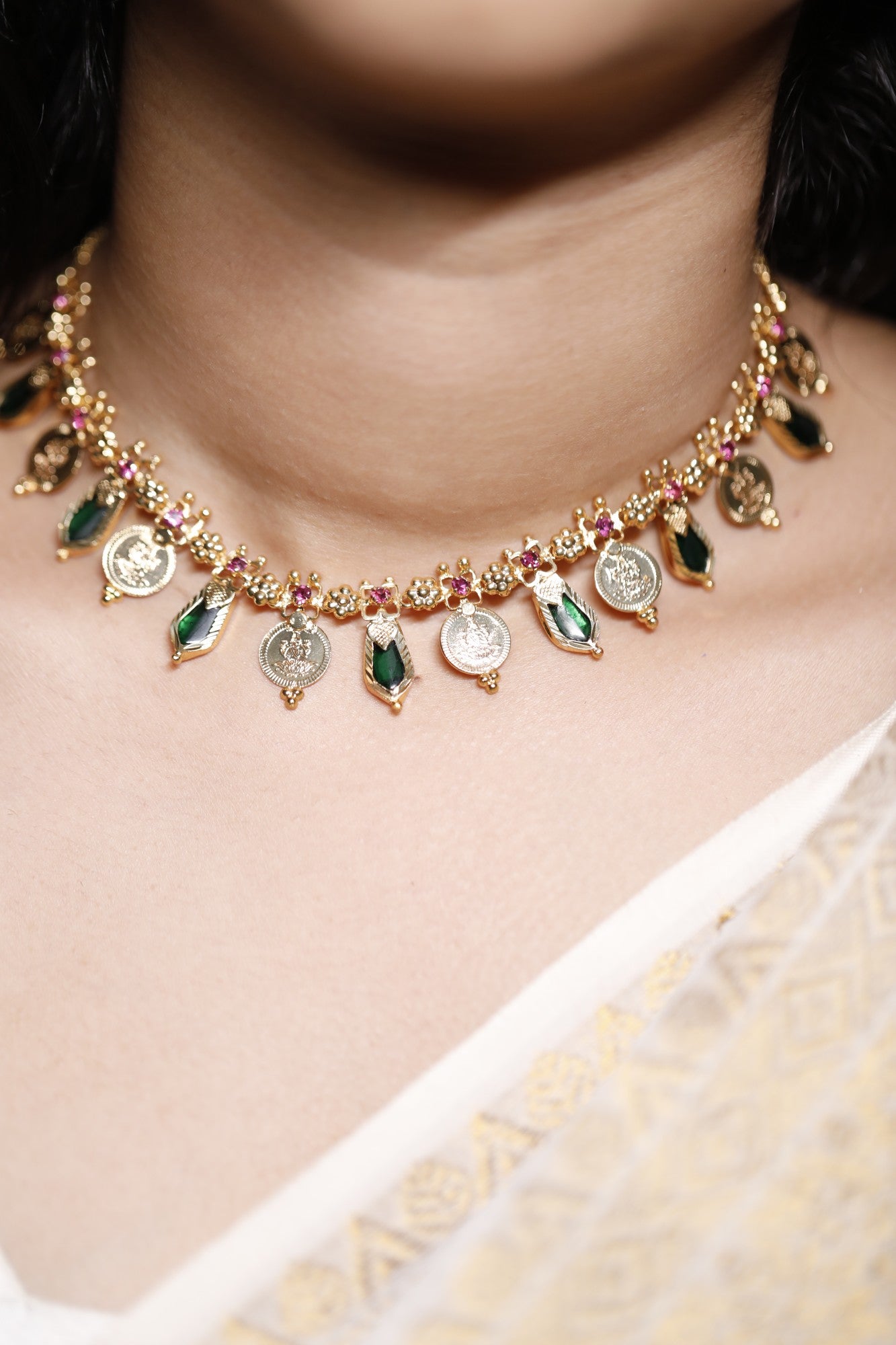 Close-up of a decorative necklace with green palakka stone and red AD with kerala traditional lekshmi idol coins and nagapadam motifs stones on a person wearing a white garment.