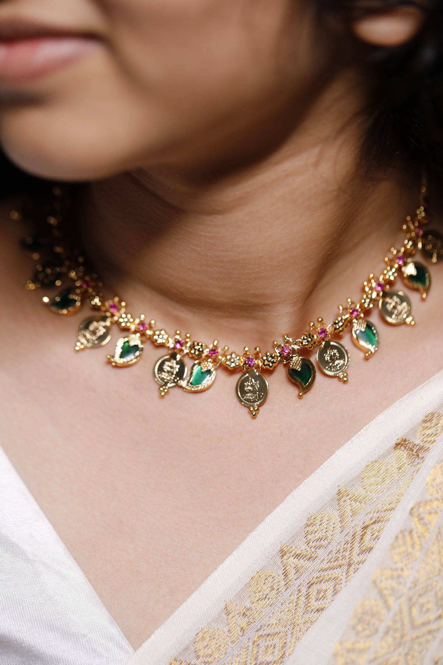 Close-up of a decorative necklace with green palakka stone and red AD with kerala traditional lekshmi idol coins and Mango motifs stones on a person wearing a white garment.