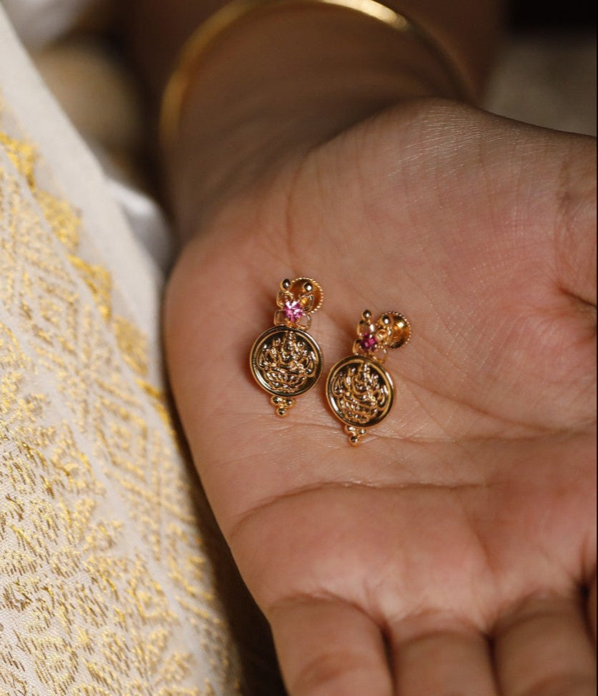 Gold Plated Lekshmi Coin earrings/studs with pink stones held in a hand against a textured fabric called Kerala Set Saree as background
