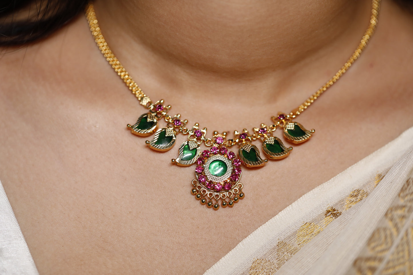 Close-up of a decorative necklace with green palakka stone and red AD stones with kerala traditional Mango motifs stones on a person wearing a white garment.