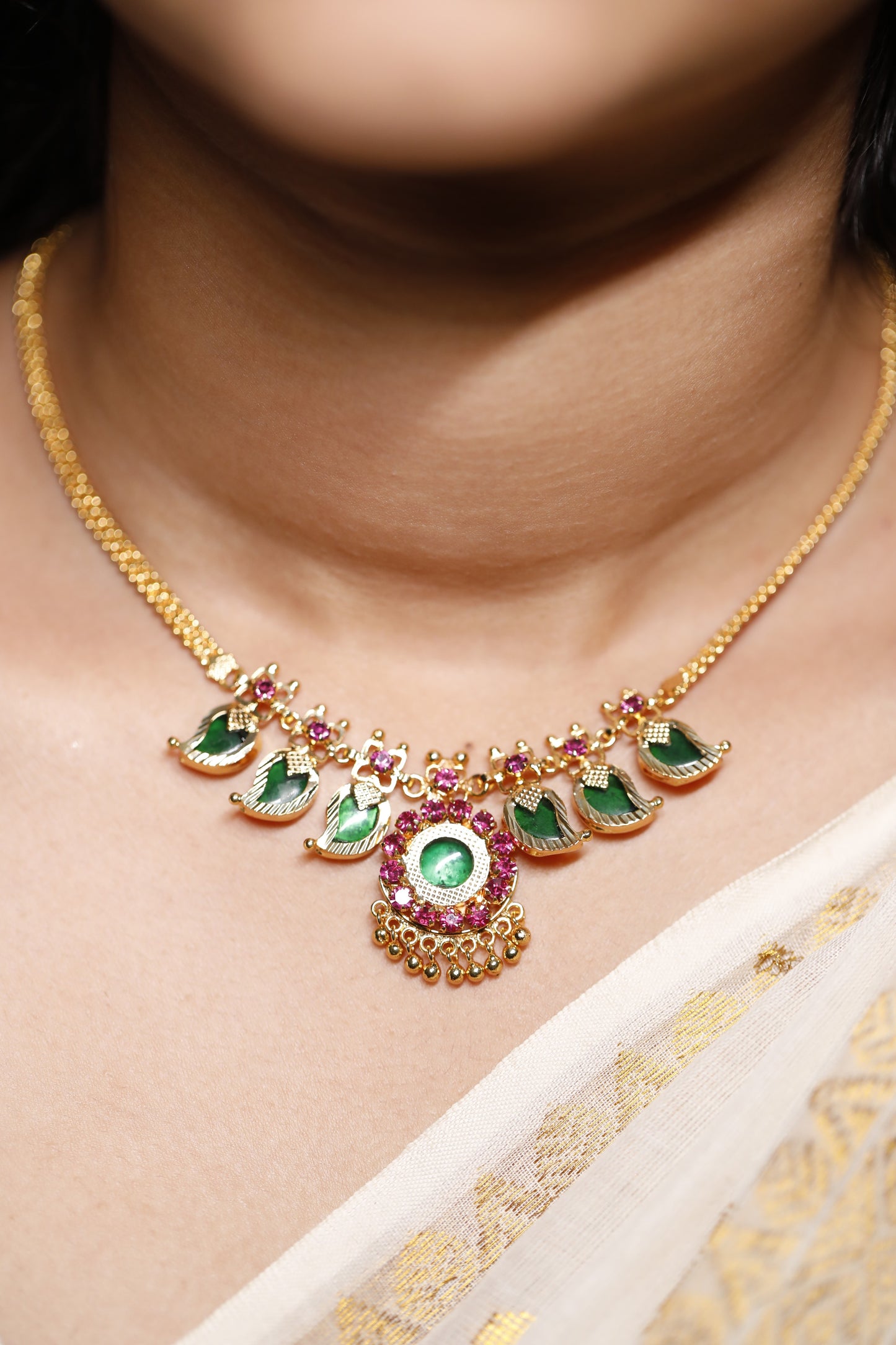 Close-up of a decorative necklace with green palakka stone and red AD stones with kerala traditional Mango motifs stones on a person wearing a white garment.