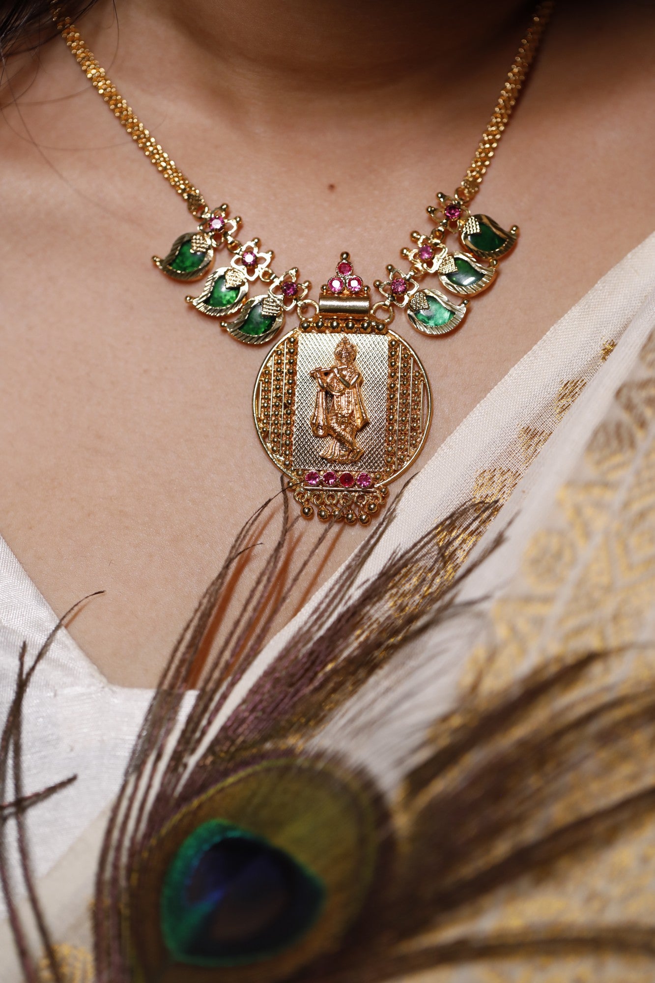 Gold plated short haram type necklace with green palakka stone and red AD stones with mango motifs and a peacock feather worn on a neck. Krishna idol in the pendant design.