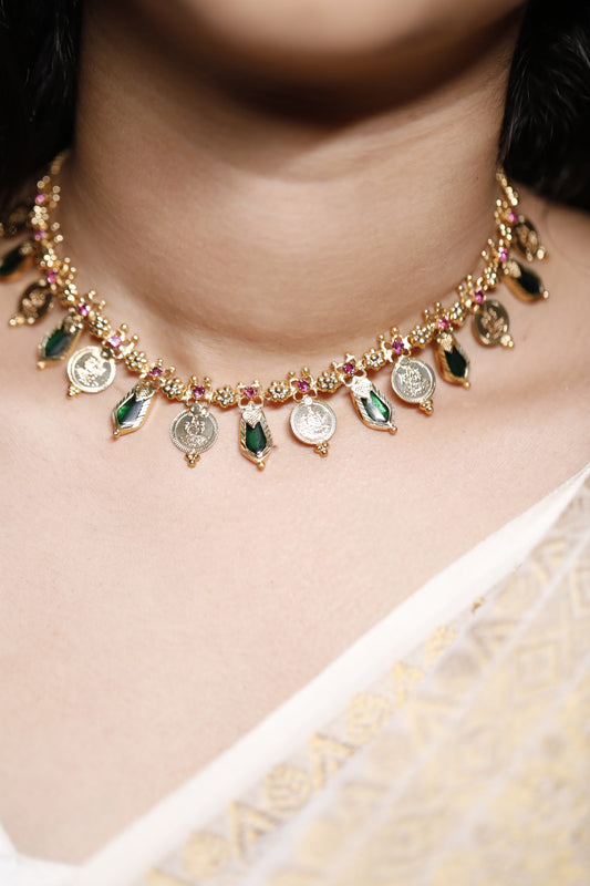 Close-up of a decorative necklace with green palakka stone and red AD with kerala traditional lekshmi idol coins and nagapadam motifs stones on a person wearing a white garment.