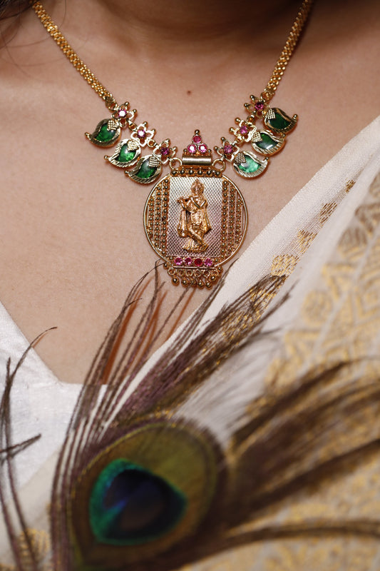 Gold plated short haram type necklace with green palakka stone and red AD stones with mango motifs and a peacock feather worn on a neck. Krishna idol in the pendant design.