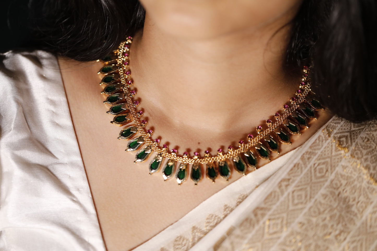 Gold plated necklace with green palakka gemstones and kerala nagapadam motifs worn by a person in a white saree.