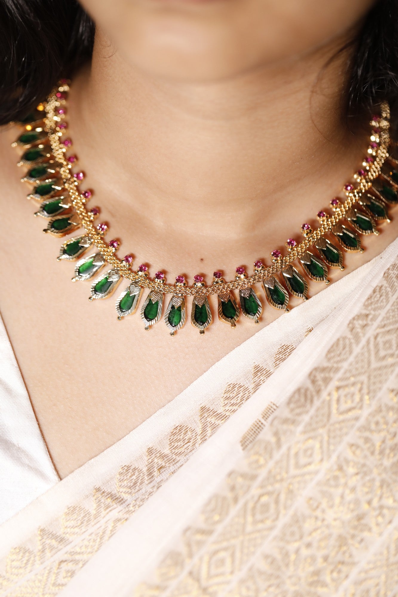 Gold plated necklace with green palakka gemstones and kerala nagapadam motifs worn by a person in a white saree.