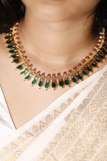 Gold plated necklace with green palakka gemstones and kerala nagapadam motifs worn by a person in a white saree.